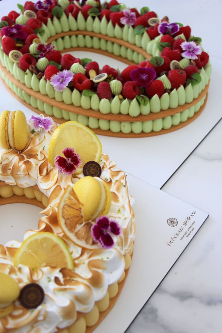 Deux Number Cakes artisanaux : un citron meringué décoré de macarons et un pistache framboise garni de framboises fraîches, pistaches et fleurs comestibles.